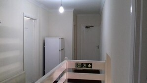 A narrow hallway with white walls, a white door, and a white radiator. There is a white refrigerator against the wall and a sign indicating a fire exit. - Haven House Hotel (Southend-on-Sea)