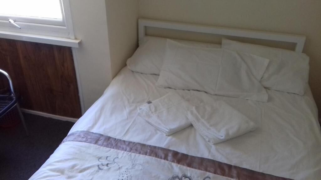 A bed with white bedding and a wooden headboard, a window with blinds, and a chair in the corner.