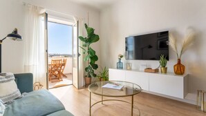 A modern living room with a sofa, a glass coffee table, a TV on a white cabinet, and a large plant. - Clifftop Nest Arrifana by Lisbeyond (Aljezur)