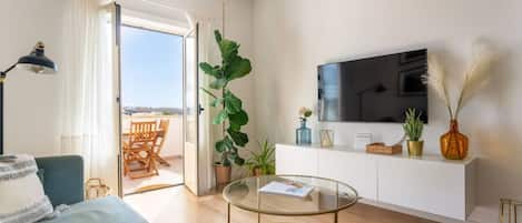 A modern living room with a sofa, a glass coffee table, a TV on a white cabinet, and a large plant.