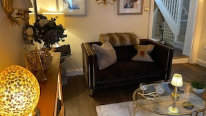 A living room with a dark velvet sofa, a glass coffee table, and a decorative lamp. There are framed pictures on the wall and a staircase in the background. - 'cosy Cottage' - 2 Bed High End Cottage in Bawtry (Doncaster)