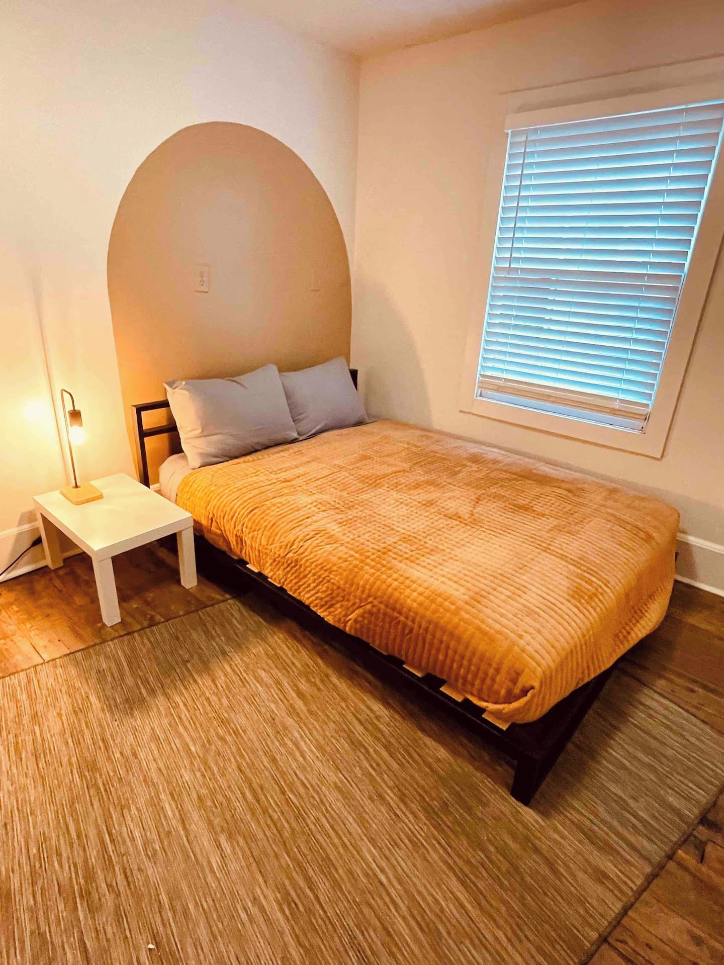 A bedroom with a bed, a nightstand, a lamp, a window with blinds, and a wooden floor.