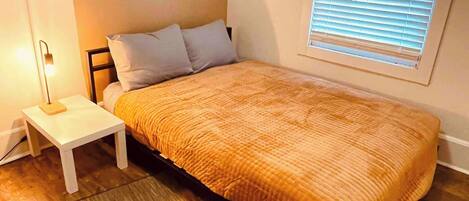 A bedroom with a bed, a nightstand, a lamp, a window with blinds, and a wooden floor.