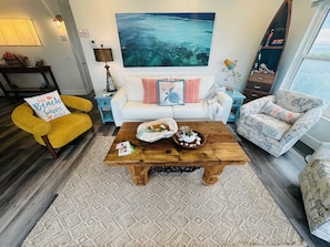 Ein Wohnzimmer mit einem weißen Sofa, einem gelben Sessel, einem Holztisch und einem großen Gemälde, das eine Strandansicht zeigt.