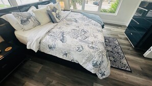 Un dormitorio con una cama grande, varios almohadones, una mesita de noche, un aparador y vistas a la playa a través de amplias ventanas.