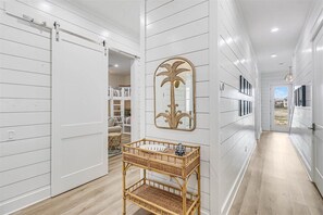 A hallway with white paneled walls, a sliding door, a mirror with a palm tree design, and a wicker console table. - Spacious, Pet-Friendly, Home w/Access to Pools &More Amenities Cinnamon Shore (Port Aransas)