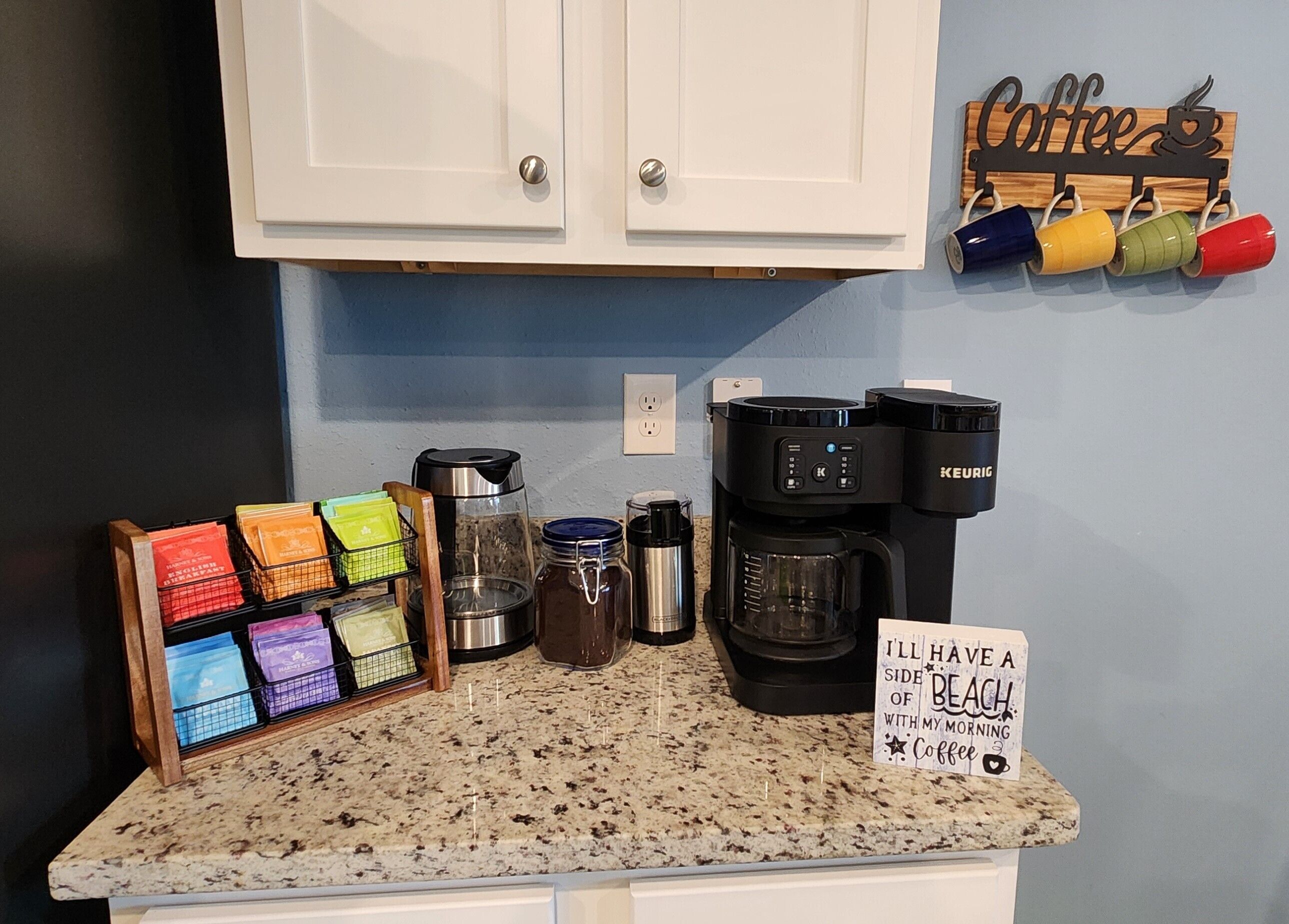 A kitchen counter with a coffee maker, mugs, and a sign that says "I'll have a side of beach with my morning coffee."
