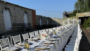 A long outdoor dining table set up with white chairs, tables, and a stone building in the background. - ROZAFA (Bujanovac)