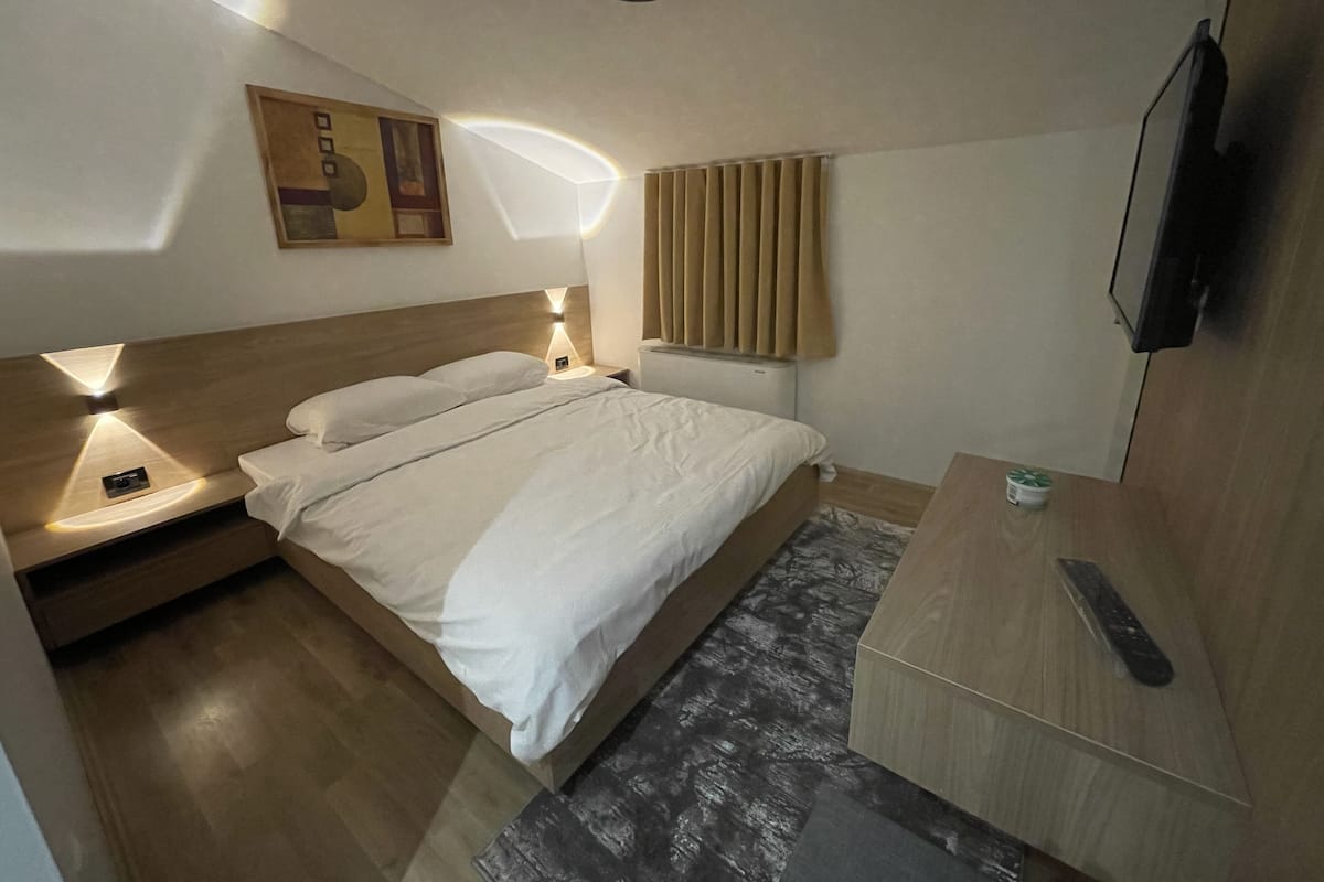 Une chambre d’hôtel moderne avec un lit en bois, des lampes de chevet, un téléviseur à écran plat et un petit bureau équipé d’une télécommande.