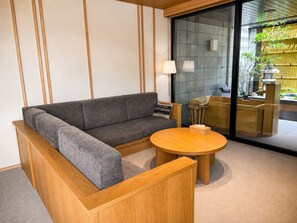 A modern interior with a sectional sofa, a round wooden table, and a glass partition with a view of a bamboo garden. - Nara Ryokan Annex Sweet Hotaru Gen (Nara)