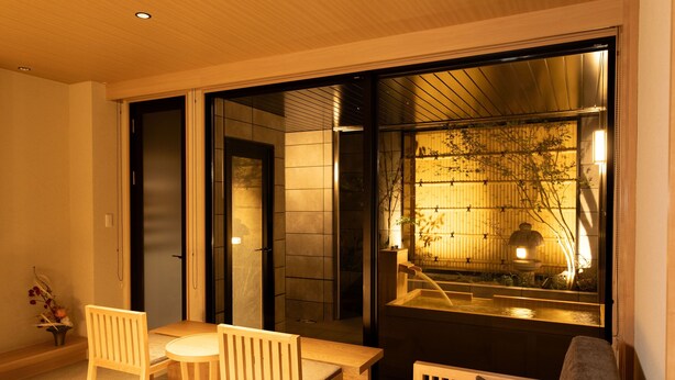 Un intérieur moderne avec une table et des chaises en bois, un canapé et une grande porte-fenêtre coulissante donnant sur un espace jardin agrémenté d’un élément d’eau et de plantes.