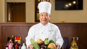 Un chef tenant une assiette de légumes dans une pièce bien éclairée, ornée d’un lustre, de boiseries et d’un téléviseur fixé au mur.