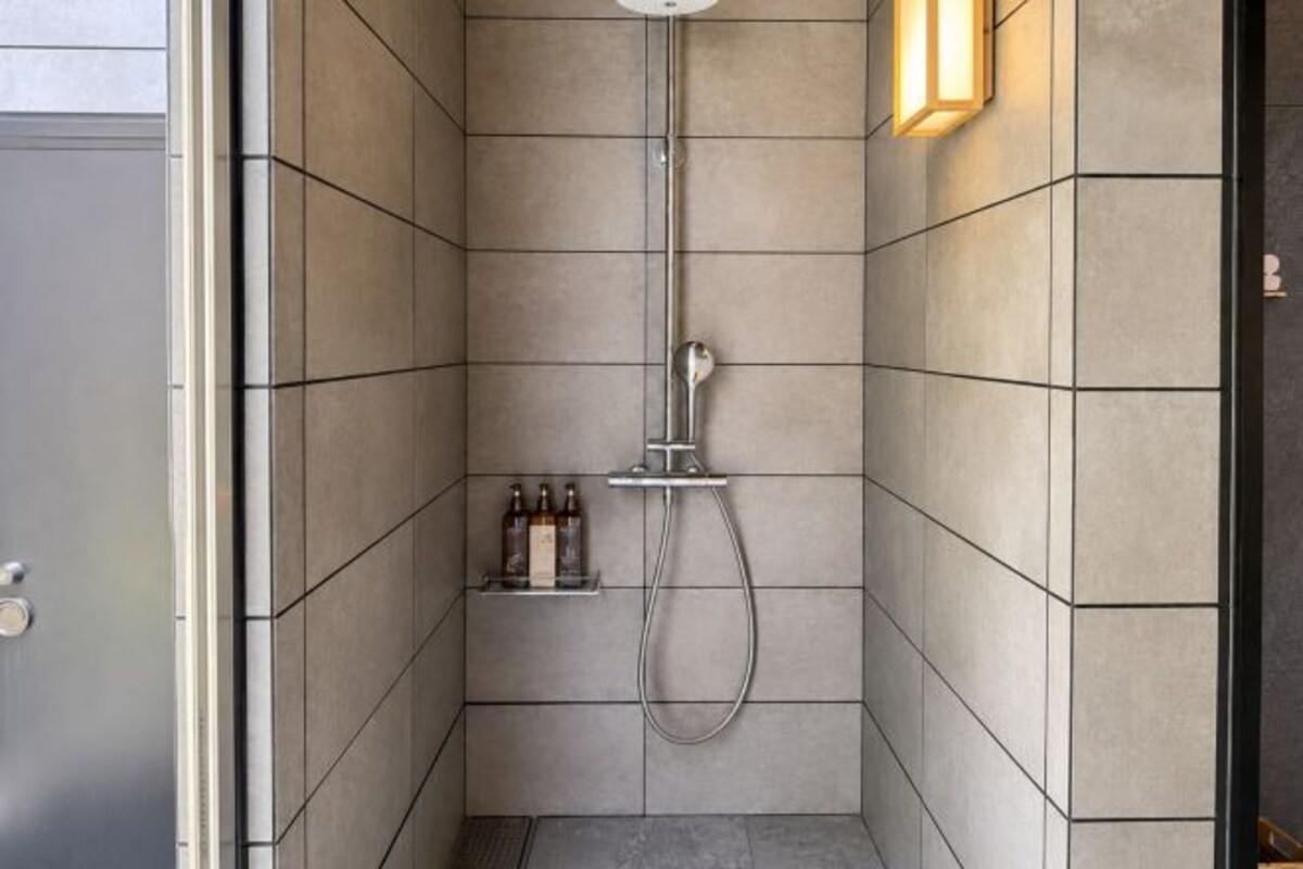 A modern shower with a handheld showerhead, a wall-mounted light, and a shelf with bottles.