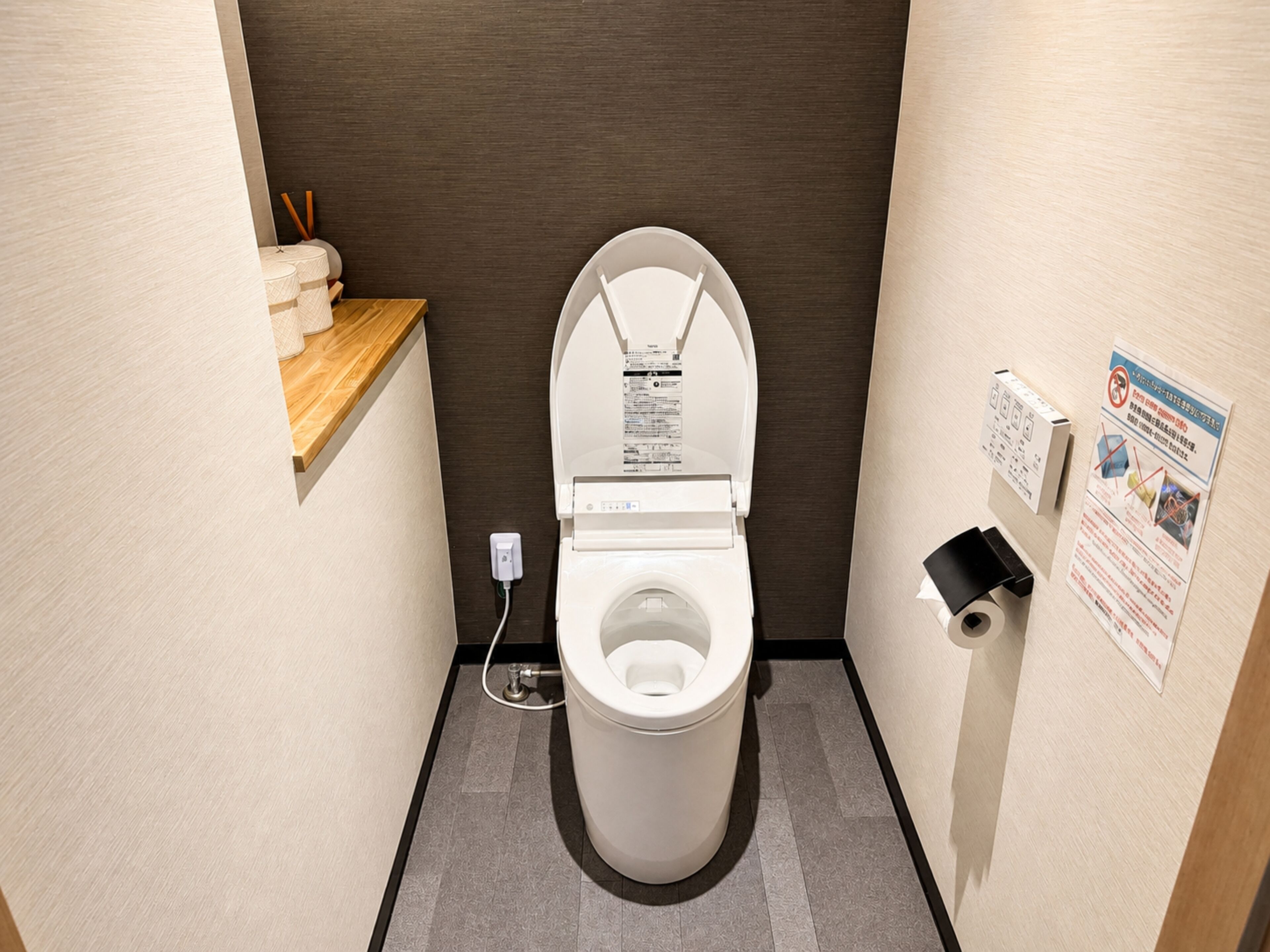A small, modern bathroom with a toilet, a wooden shelf with toiletries, a paper towel dispenser, and a safety sign on the wall.