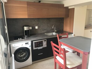 Compact kitchen with a modern design, featuring a washing machine, coffee maker, and microwave, and a small dining area with a table and two chairs.