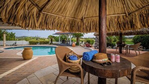 Ein Poolbereich mit einem Reetdachschirm, Korbstühlen, einem Tisch mit Hüten und Dosen und Blick auf den Pool und die Landschaft.