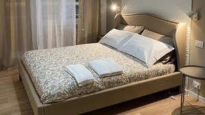 Una cama bien hecha con una colcha estampada y almohadas, una mesita de noche con una lámpara, y una ventana con cortinas de encaje.