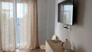 Una habitación moderna con un escritorio de madera, un televisor montado en la pared y una puerta corrediza de vidrio con cortinas color beige.