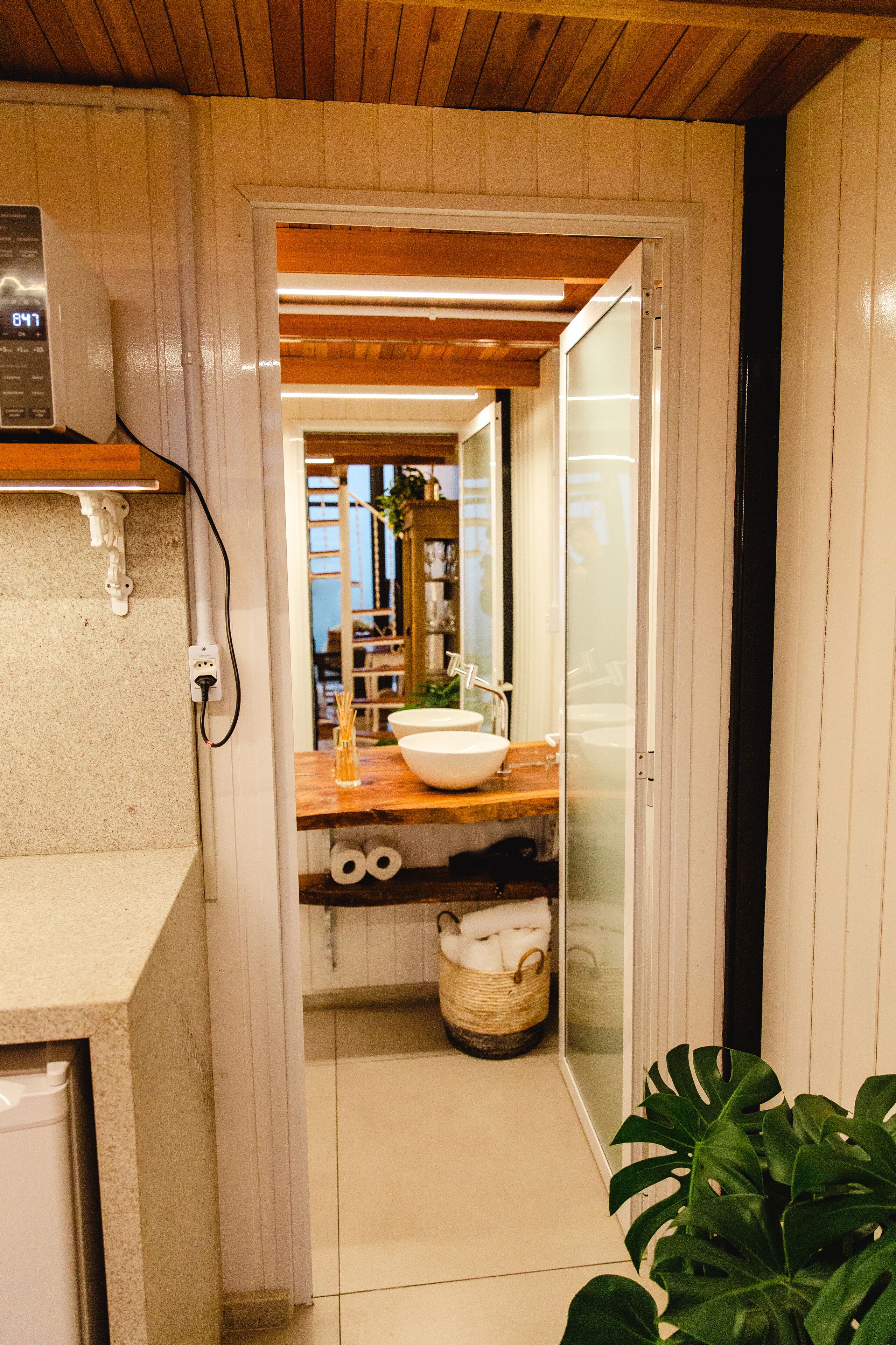 Un couloir étroit mène à une salle de bain dotée d’un plafond en bois, d’un lavabo blanc et d’un miroir.