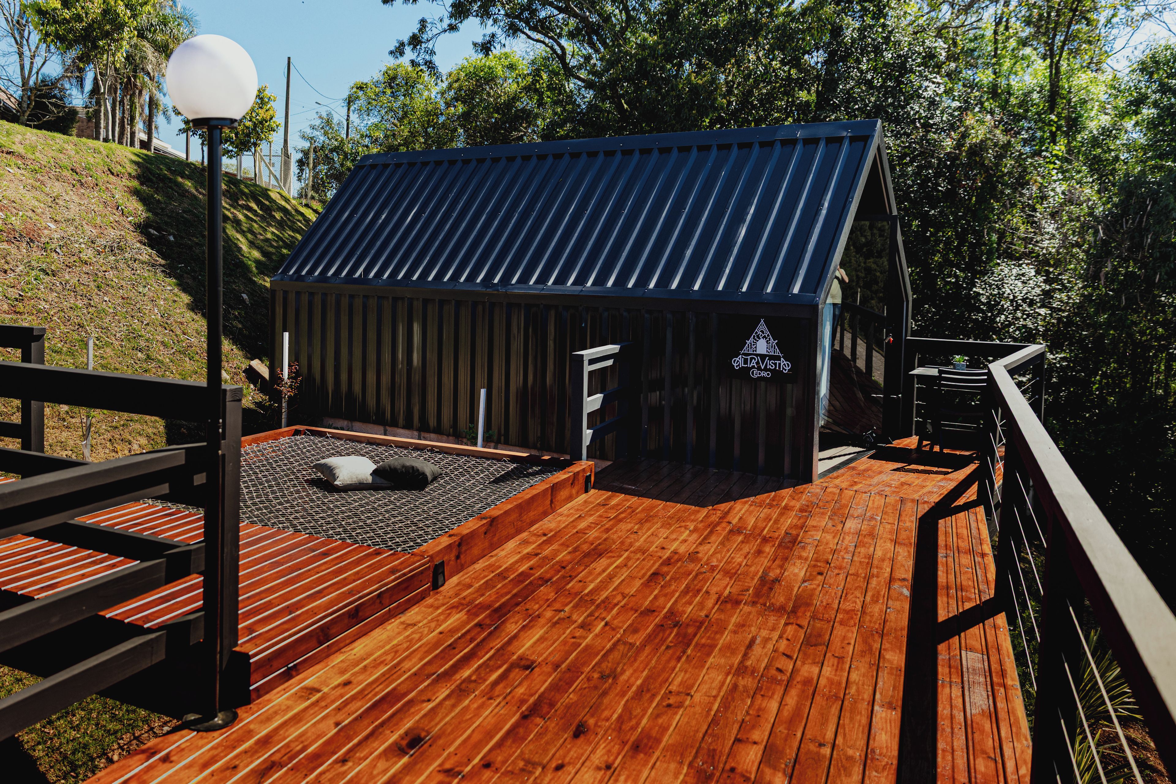 Una terraza de madera con una estructura de techo metálico, posiblemente una sauna o cocina al aire libre, rodeada por una valla de madera y un farol.