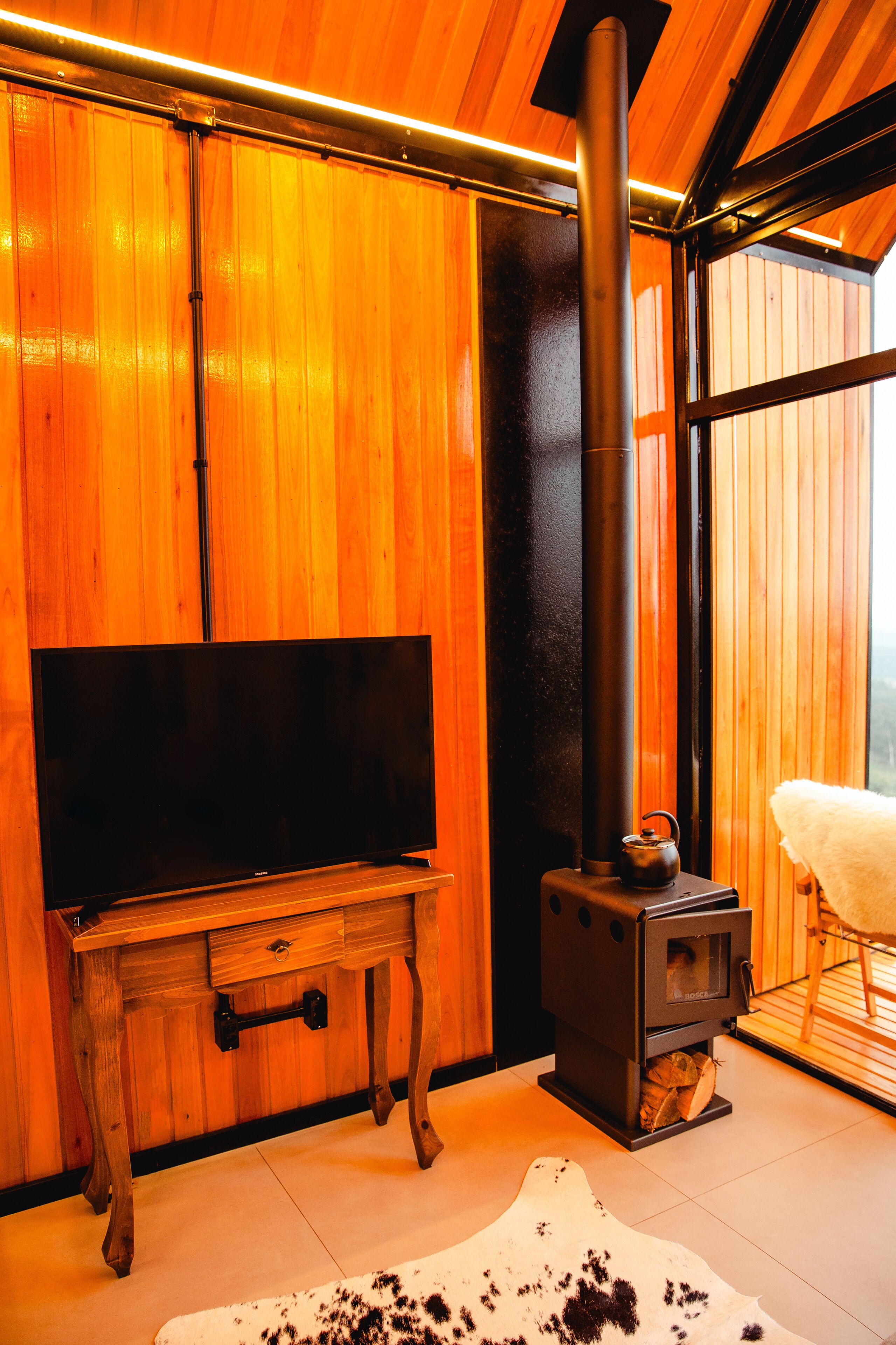 Un interior de madera con televisor, estufa de leña y un ventanal grande.