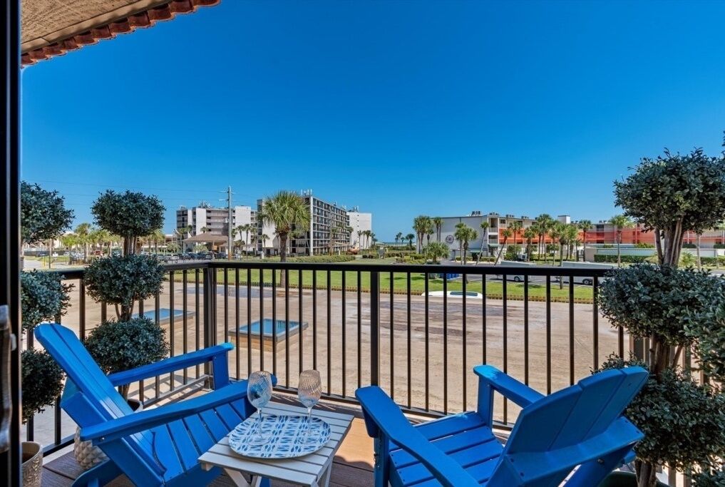 A balcony with blue chairs and a table, overlooking a parking area and buildings.