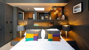 Ein modernes Schlafzimmer mit einem großen Bett, zwei Nachttischen mit Lampen, einer eingebauten Sitzbank mit Stauraum, einem Spiegel und einer kleinen Küchenzeile.