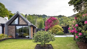 Ein modernes Haus mit Satteldach, großen Glasfenstern und einem gepflegten Garten mit blühendem Buschwerk.