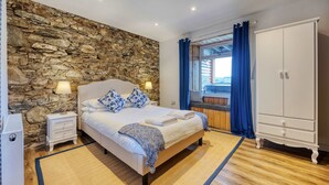 Un dormitorio con pared de piedra, una cama con almohadas, una mesita de noche, un armario y una ventana con cortinas.