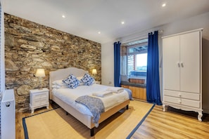 A bedroom with a stone wall, a bed with pillows, a nightstand, a wardrobe, and a window with curtains. - Blar Cottage (Private Sauna + Fast Starlink Wifi) (Ardbrecknish)