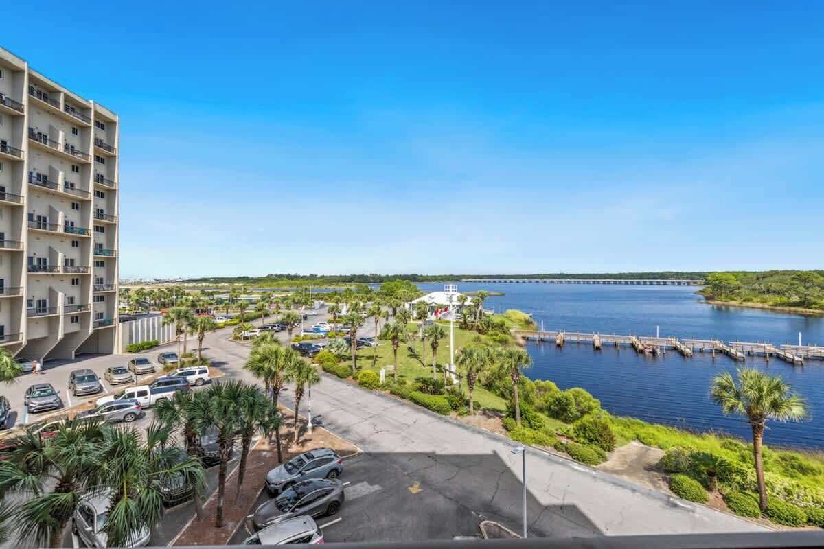 A high-rise hotel with a parking lot, palm trees, and a view of a body of water with a bridge in the distance.