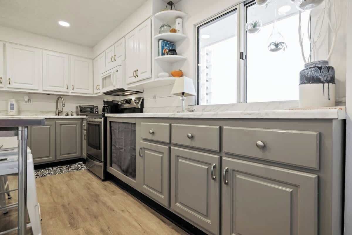 A modern kitchen with grey cabinets, a built-in oven, and a large window with decorative hanging plants.