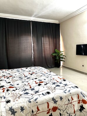 Une chambre à coucher avec un grand lit, des rideaux sombres, une plante en pot et un téléviseur à écran plat fixé au mur.