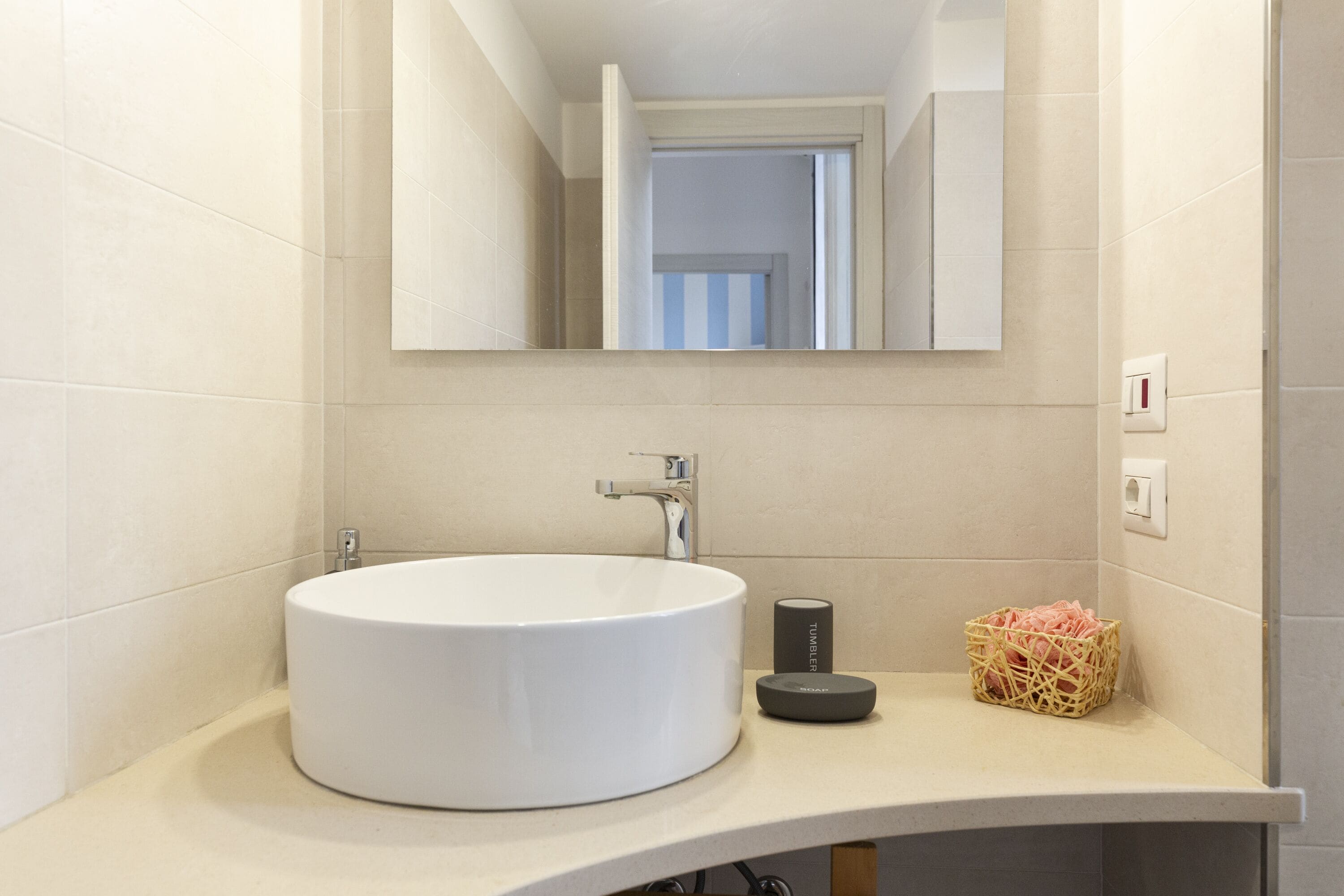 A bathroom with a round white sink, a mirror, and a small basket of pink flowers.