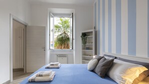 A bedroom with a large bed, a window with a view of palm trees, a bookshelf, and a door. - nota-Mi - Mezzofanti TRE PONTI (Milan)
