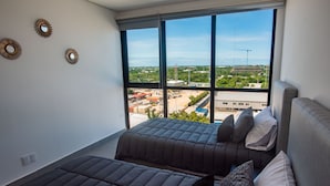 A modern bedroom with two beds, large windows, and a view of the cityscape. - New Deluxe Unit, Signature Condominium by The Harbor (Mérida)