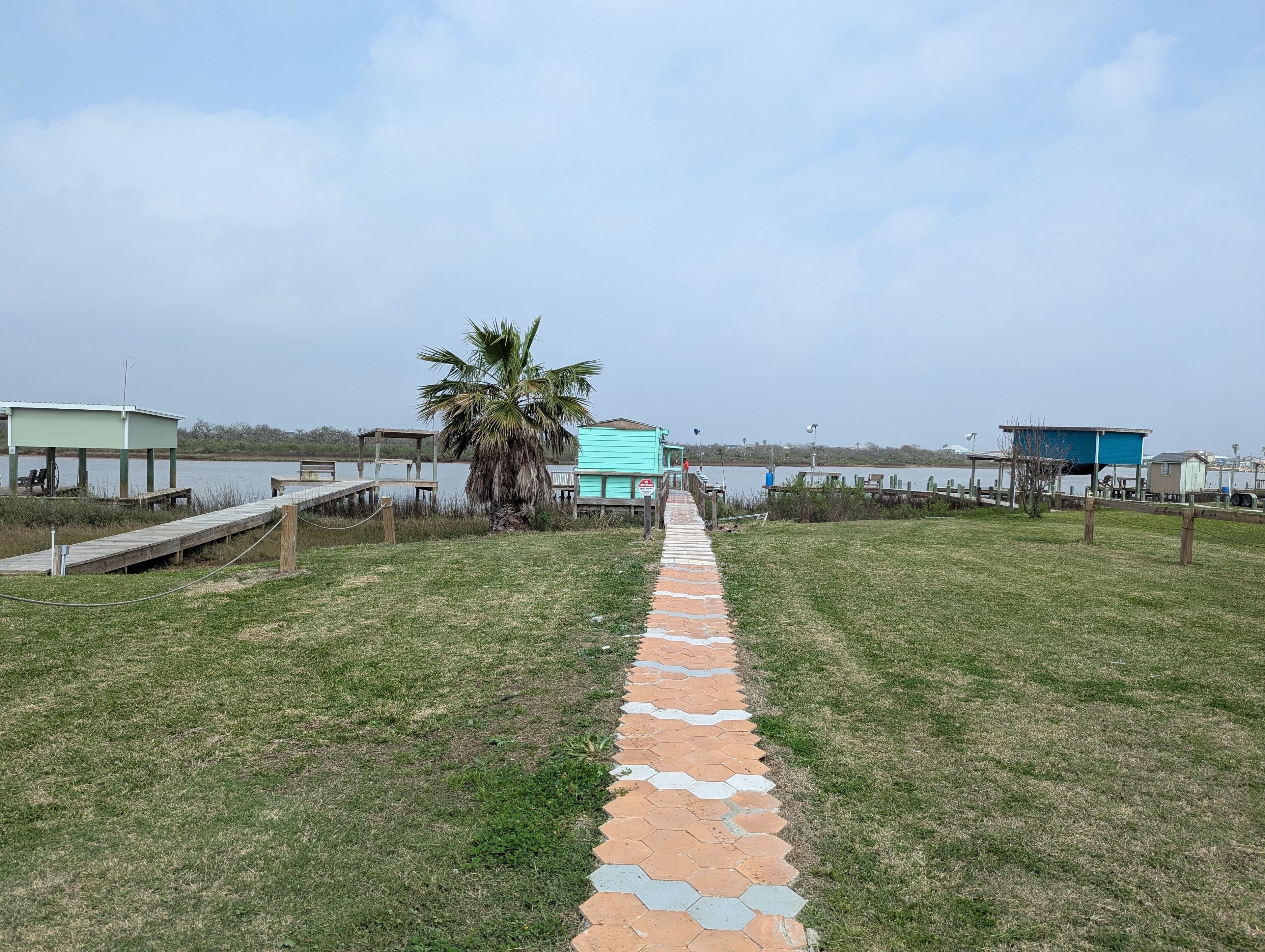 Un sendero que conduce a una zona de playa con una palmera, un pabellón y un pequeño edificio azul.