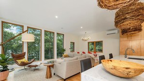 Een moderne woonkamer met grote ramen, een bank, een fauteuil, een houten schaal op een marmeren aanrechtblad en een hanglamp.