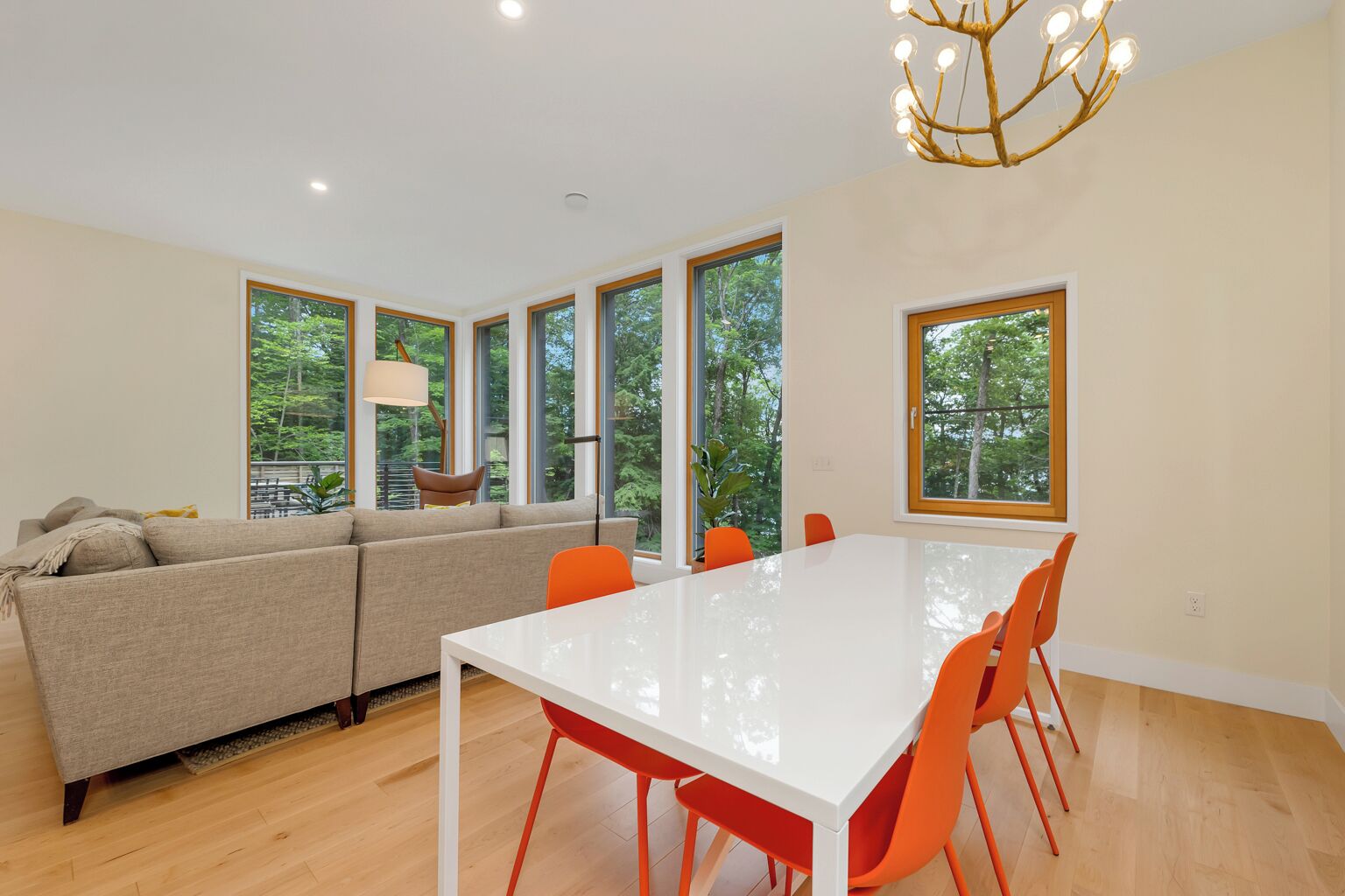 A modern living room with a white dining table, orange chairs, a beige sofa, and large windows offering a view of trees.