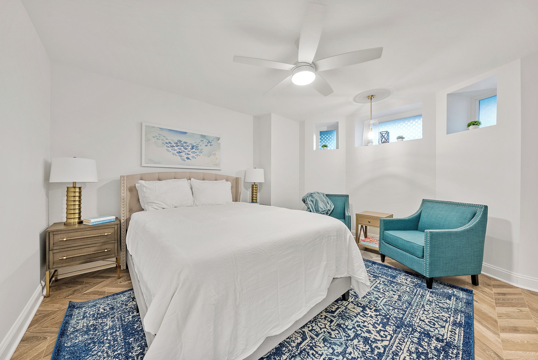 Slip into crisp white linens and let the ceiling fan's gentle breeze lull you to sleep in this serene bedroom retreat, where calming blue accents and natural light create your perfect escape.