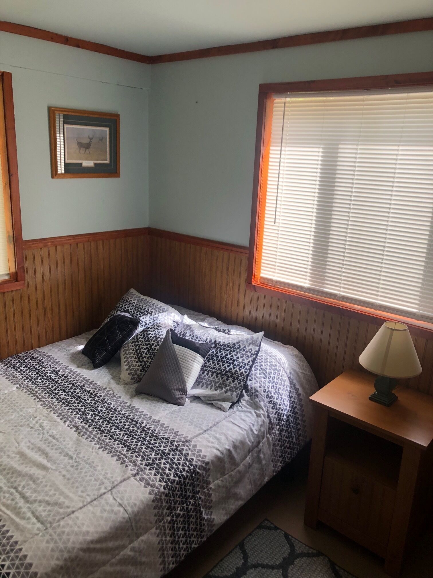 A bedroom with a bed, a nightstand, a lamp, a window with blinds, and a framed picture on the wall.