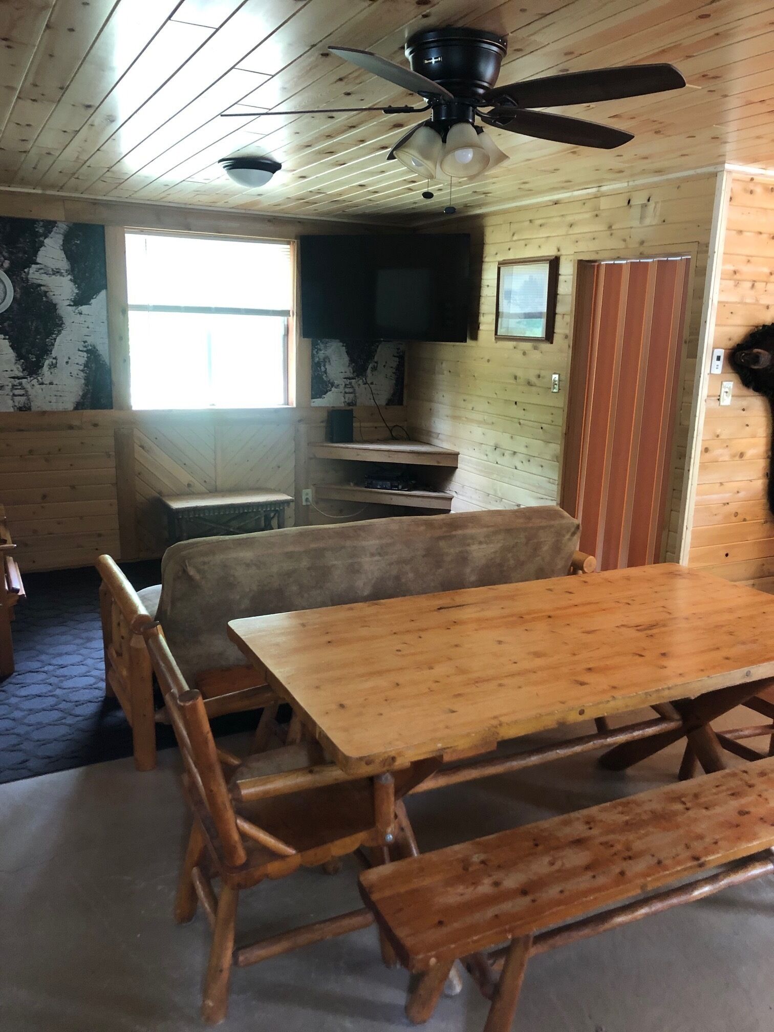 A wooden interior with a dining table, benches, a ceiling fan, and a television mounted on the wall.