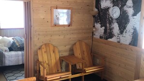 Une pièce chaleureuse de style cabane, avec des murs en bois, un ventilateur de plafond, une fenêtre avec des rideaux, un lit recouvert d’une courtepointe à motifs, deux chaises en bois, une petite table et une horloge murale.