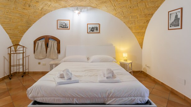 A bedroom with a brick ceiling, a bed with white linens, two framed pictures on the wall, and a nightstand with a lamp.