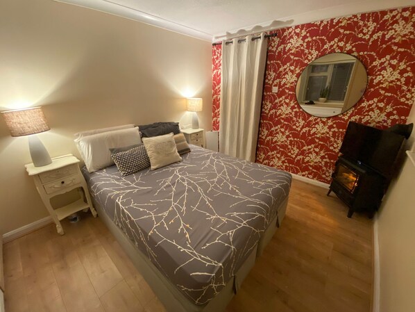 Un dormitorio con cama, dos mesitas de noche con lámparas, cómoda, espejo, calentador y papel tapiz floral.