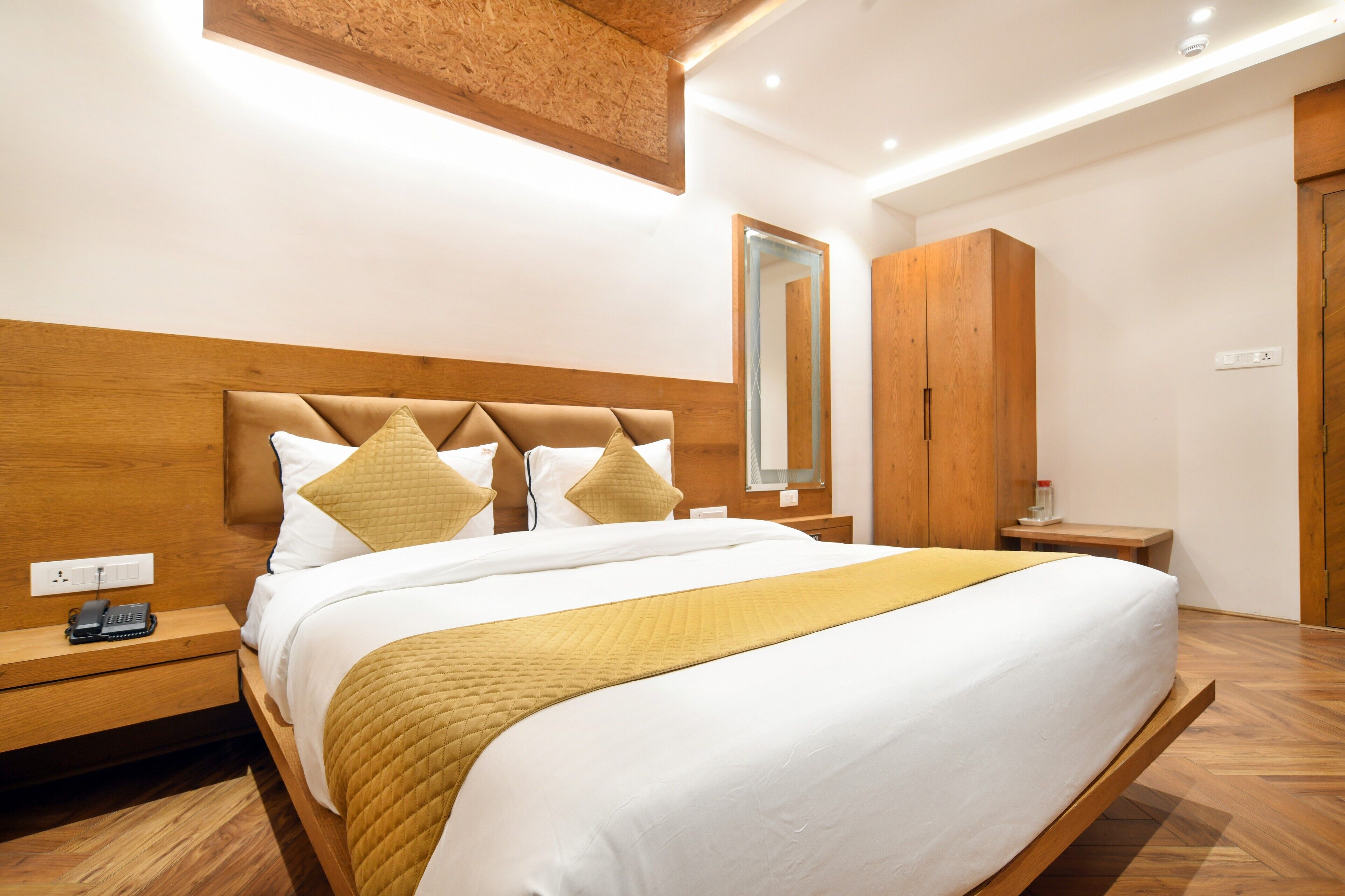 A modern hotel room with a wooden headboard, a large bed with white bedding and a yellow throw, a bedside table with a phone, and a mirror on the wall.