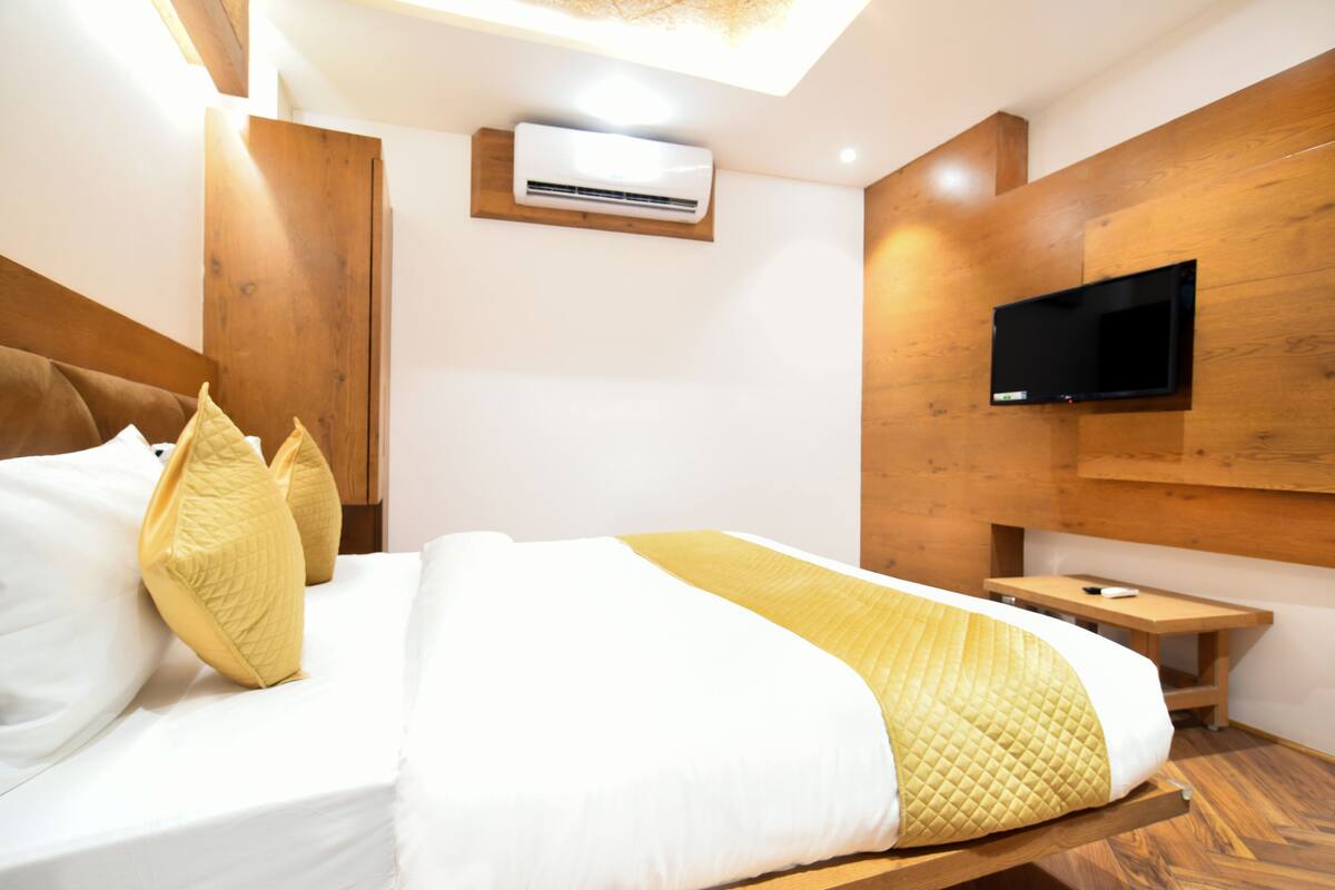 Una habitación de hotel moderna con cabecera de madera, un televisor de pantalla plana montado en la pared, una mesita de noche con control remoto y una cama con ropa blanca y una manta amarilla.