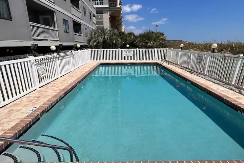Une piscine rectangulaire, entourée d’une clôture blanche et dotée de marches permettant d’y accéder, est bordée d’un bâtiment avec des balcons, et le ciel est dégagé.