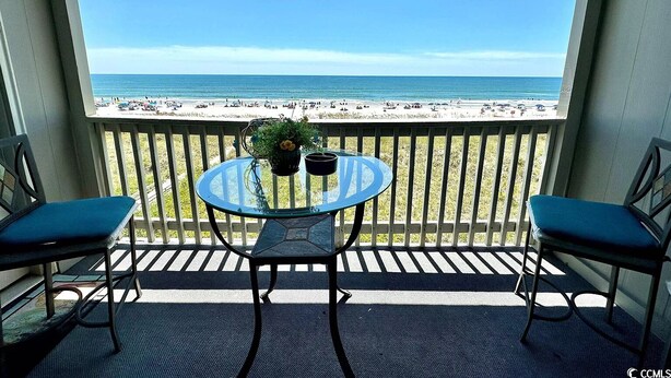 A balcony with a glass table and two chairs, overlooking a beach with people and umbrellas. - Tequila Sunrise - 2BR Oceanfront (North Myrtle Beach)