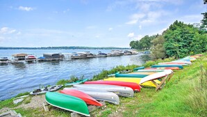 A row of colorful canoes lined up on a grassy shore by a calm body of water with boats docked in the background. - Lakeside Retreat in Dewittville w/ Pool Access! (Dewittville)